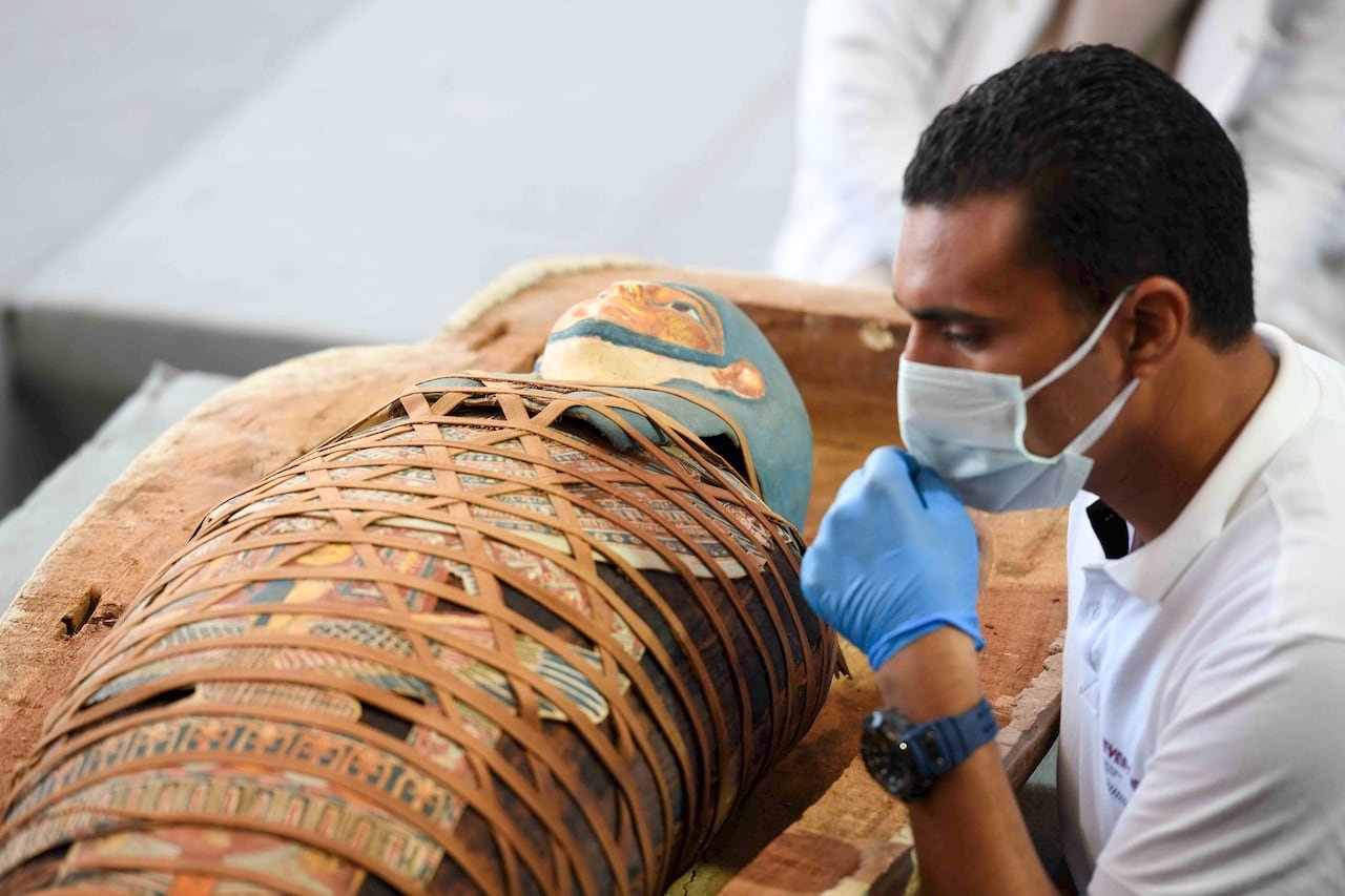 default-290 An archaeologist inspects an Egyptian mummy that is wrapped in an elaborate and colourful burial shroud. The archaeologist is wearing a medical mask and gloves.