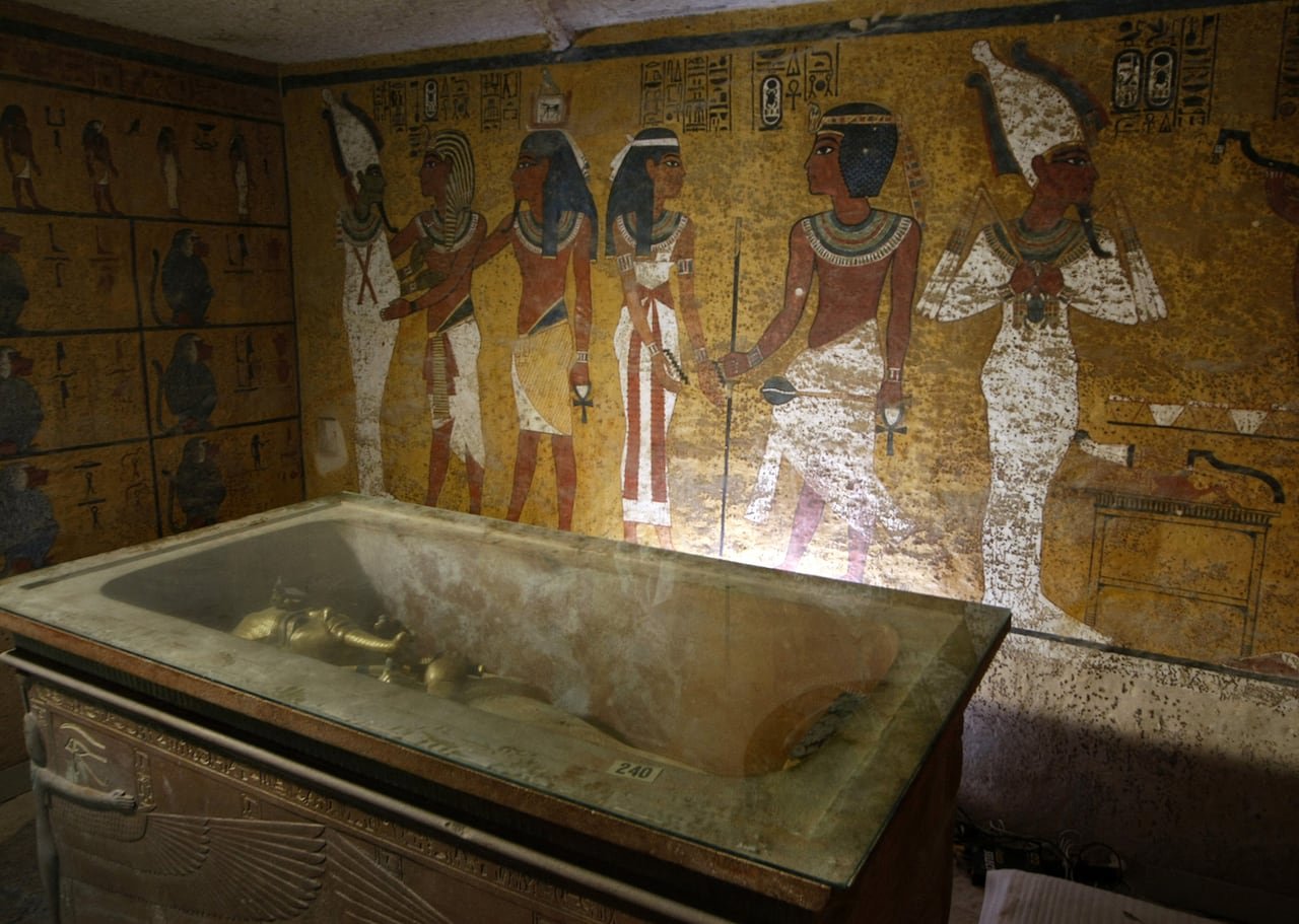 default-291 A golden sarcophagus is shown lying in an elaborate burial chamber. The wall behind it is colourfully painted with figures.